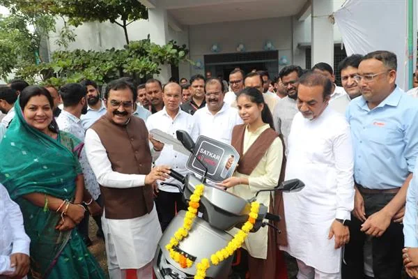 फूलों की माला से सजी स्कूटी के पास खड़ी छात्रा को चाबी सौंपते मंत्री और आसपास मौजूद लोग