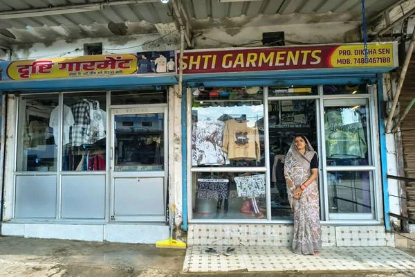 कपड़ों की दुकान के बाहर साड़ी पहने खड़ी महिला, पीछे शीशे की अलमारियों में वस्त्र दिखाई दे रहे हैं