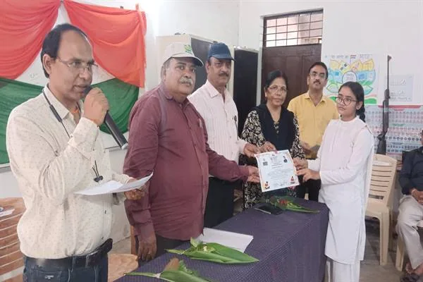 विद्यालय के कक्ष में मंच के पास एक छात्रा को प्रमाणपत्र देते शिक्षक और अतिथि, सामने मेज पर फूल रखे हैं