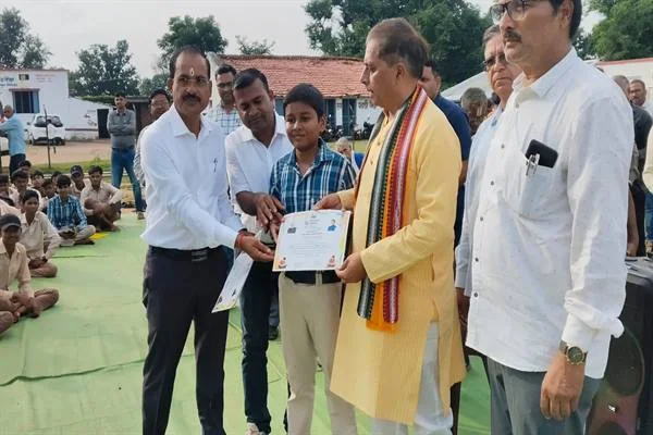 खुले परिसर में पीले कुर्ते पहने व्यक्ति और अन्य लोग एक छात्रा को प्रशस्ति-पत्र देते हुए, पीछे बैठे विद्यार्थी