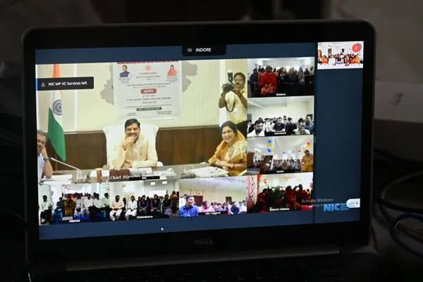 लैपटॉप की स्क्रीन पर चल रही वर्चुअल कॉन्फ्रेंस, जिसमें मंच पर बैठे दो लोग दिख रहे हैं और किनारे कई जिलों के कार्यक्रम स्थलों की छोटी-छोटी विंडो में छात्र-छात्राएं नजर आ रहे हैं।