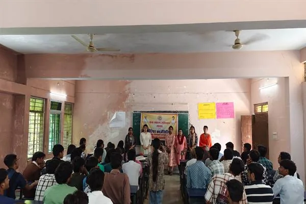 कक्षा-कक्ष में बेंचों पर बैठे विद्यार्थियों के सामने खड़ी कुछ महिलाएं और छात्राएं, पीछे दीवार पर हरे रंग का बैनर टंगा है।