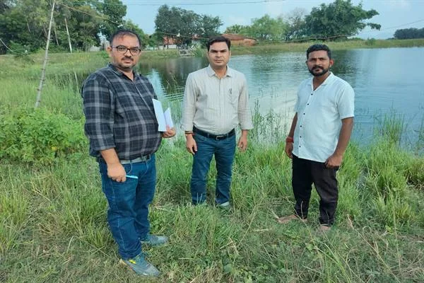 एक बड़े तालाब के किनारे घास पर खड़े तीन पुरुष, पीछे पानी, झाड़ियाँ और पेड़ दिख रहे हैं; बाएं खड़े व्यक्ति के हाथ में कागज हैं।