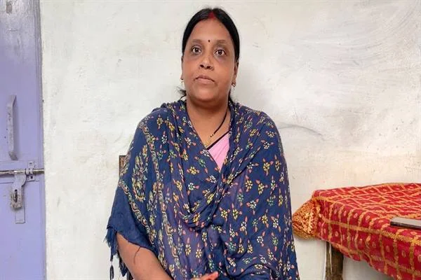 नीली बूटीदार साड़ी और दुपट्टे में एक महिला घर के भीतर सफेद दीवार के सामने बैठी हैं; पीछे बैंगनी दरवाजा और लाल-सुनहरे कपड़े से ढकी मेज दिखाई दे रही है।