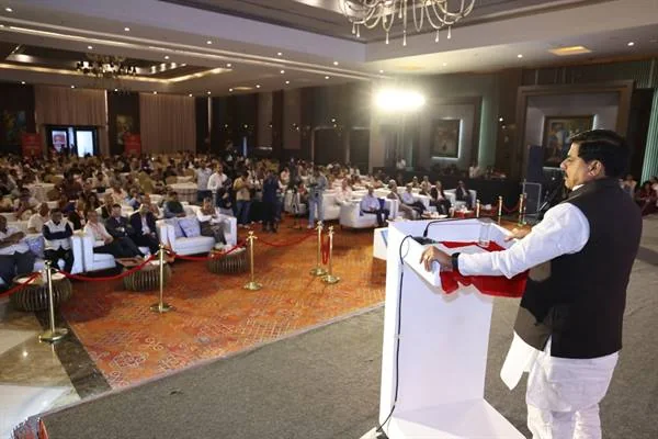 होटल के हॉल में मंच पर सफेद डायस के पीछे खड़े होकर माइक पर संबोधित करते मुख्यमंत्री, सामने कुर्सियों पर बैठे बड़ी संख्या में श्रोता।